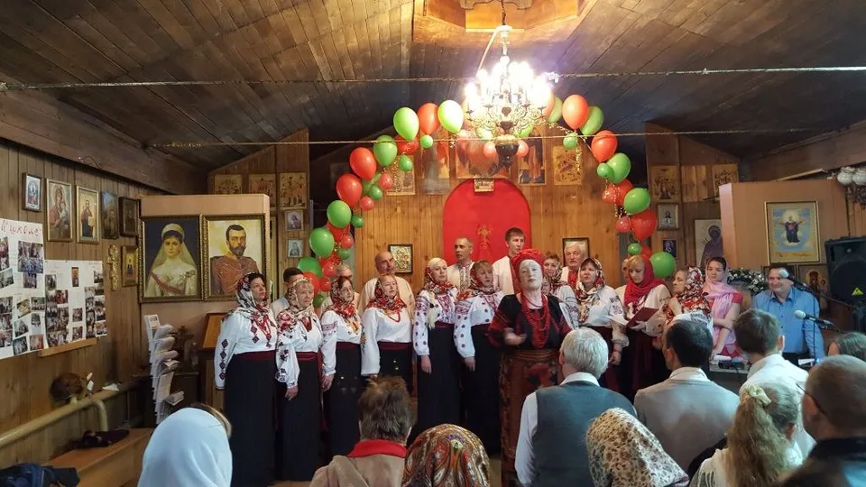 Хор в народных костюмах поёт в небольшом деревянном зале, украшенном воздушными шарами и образами святых Кирилла и Мефодия