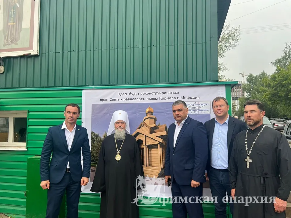 Группа людей, включая архиерея и чиновников, стоит у зелёного строительного вагончика с баннером о реконструкции храма