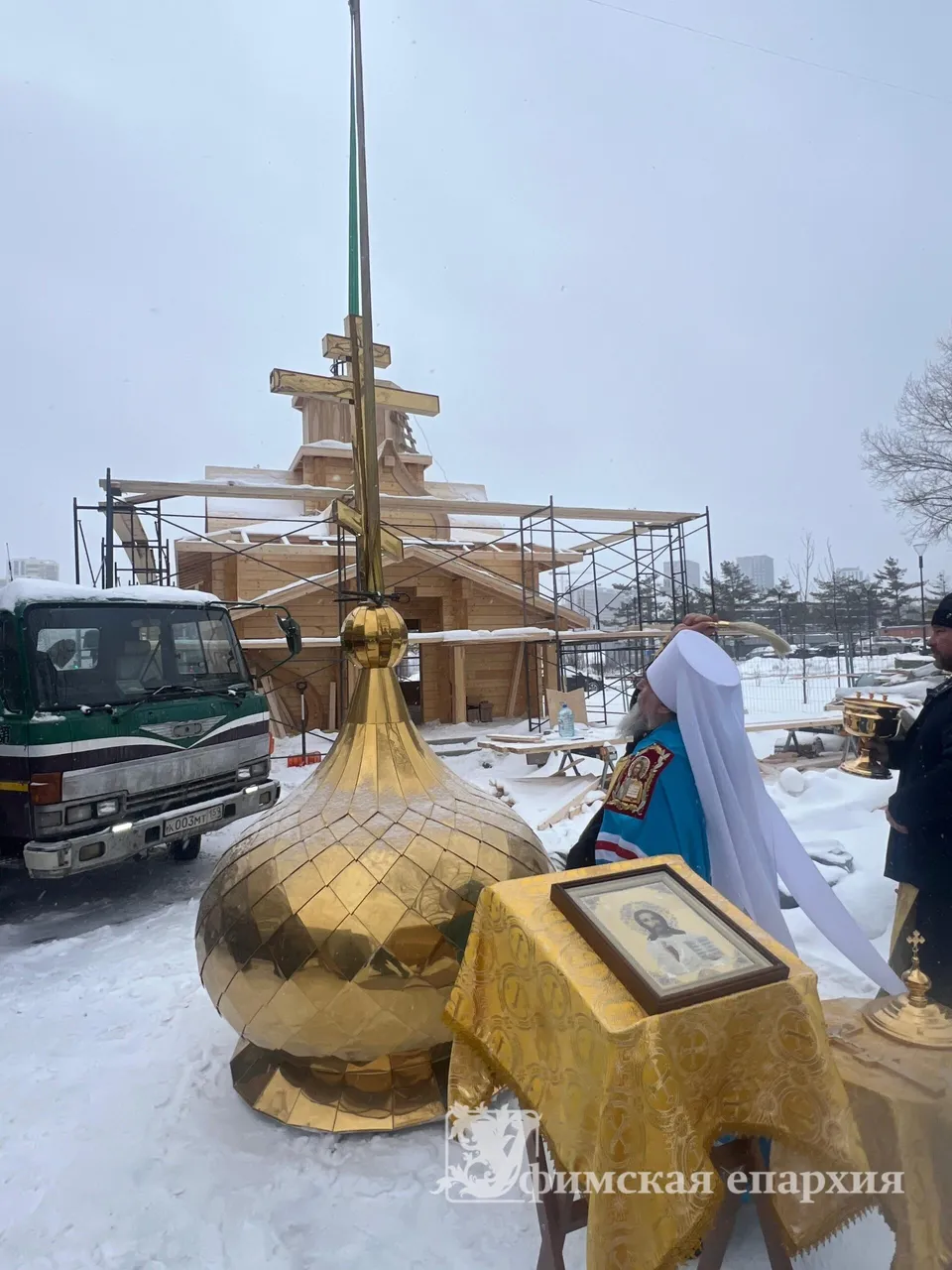 Золотой купол с крестом на кране готовят поднять на крышу деревянного храма, рядом архиерей совершает освящение